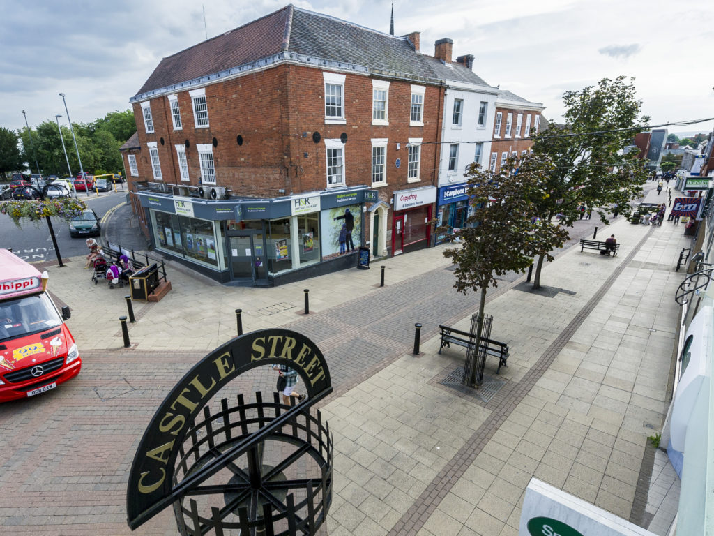 Image of the Castle Street Hinckley & Rugby branch from a slight distance with a view of the high street containing trees, signs and an ice-cream van.