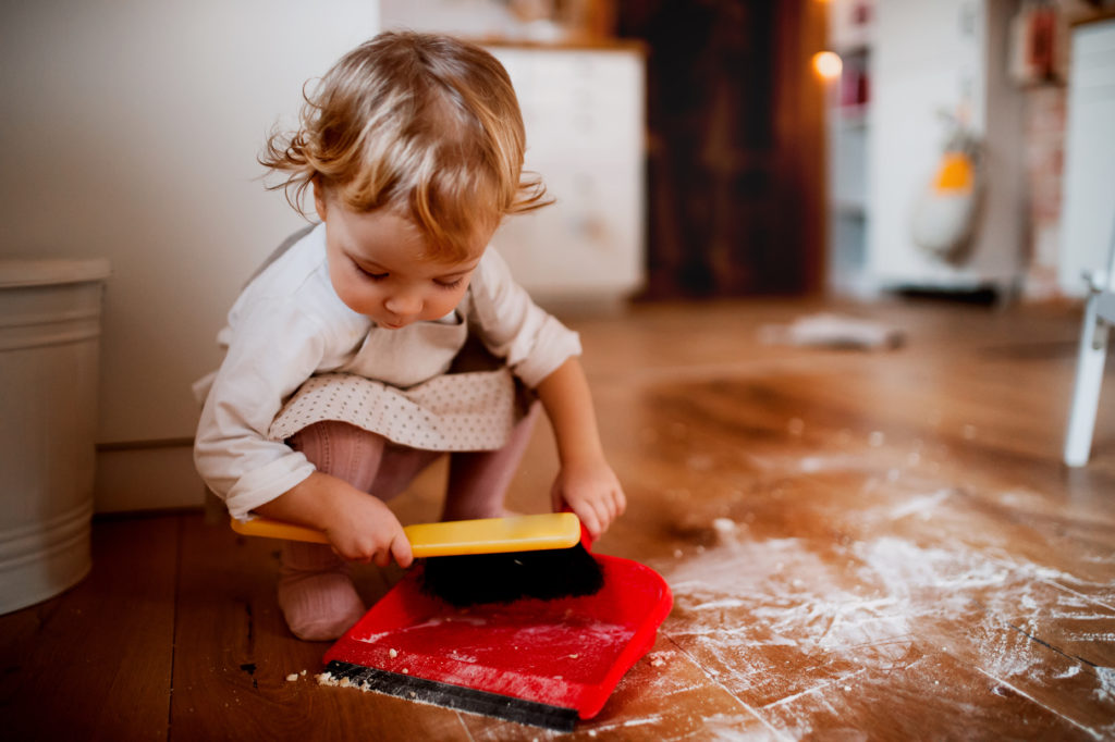 Little toddler with a dustpan and brush trying to clean the flour on the floor.