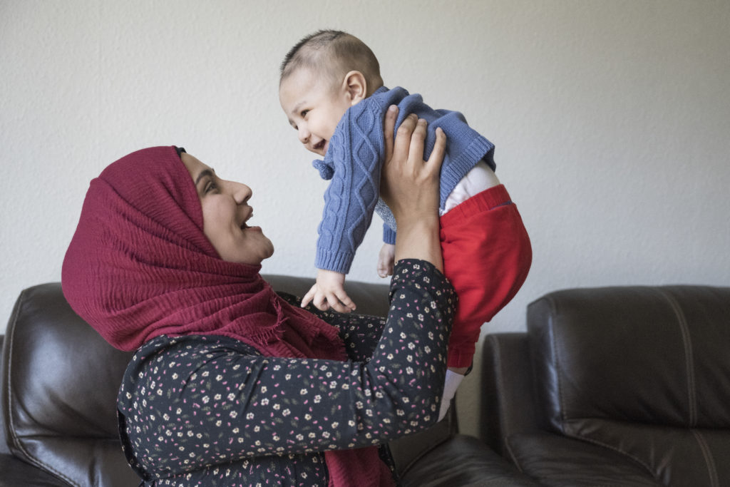 Side view of mid adult woman in headscarf and tunic face to face with her laughing 5 month old son as they enjoy time together in living room of family home.