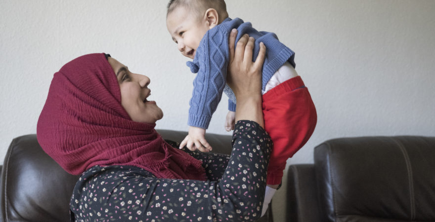 Side view of mid adult woman in headscarf and tunic face to face with her laughing 5 month old son as they enjoy time together in living room of family home.