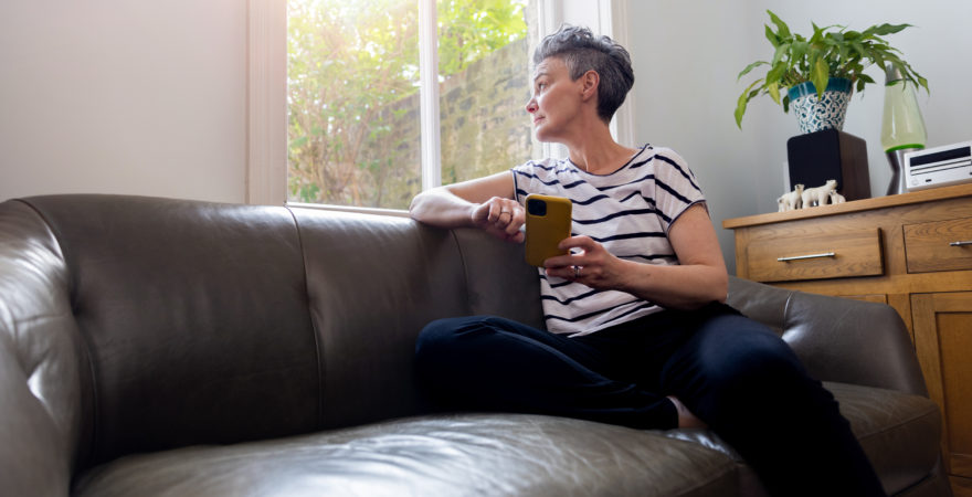 A three quarter length shot of a mature female adult sitting on a leather sofa. She has her phone in her hands whilst looking out the window.