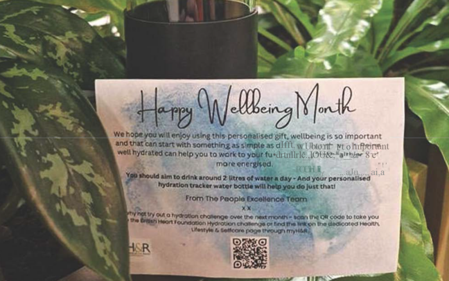 Image of a personalised note titled happy wellbeing month with green leaves in the background and a glass water bottle.