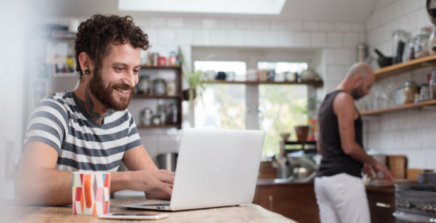 A couple hanging out in their kitchen with one of them sitting at a breakfast table smiling at their laptop.
