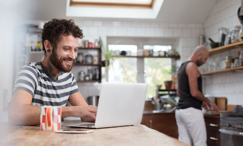 A couple hanging out in their kitchen with one of them sitting at a breakfast table smiling at their laptop. 