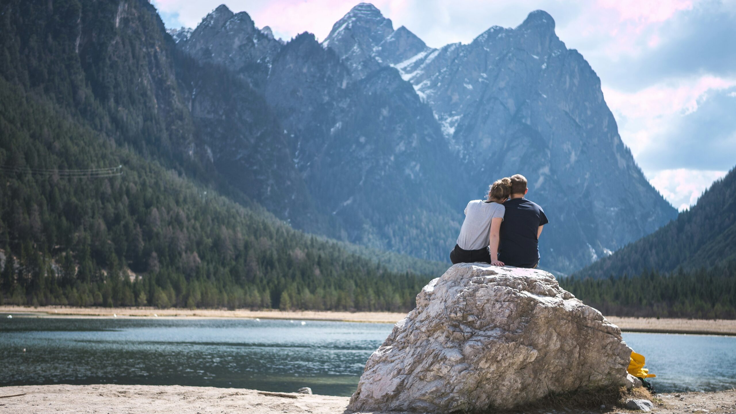 A couple sat on a rock looking out at a lake with mountains in the distance.