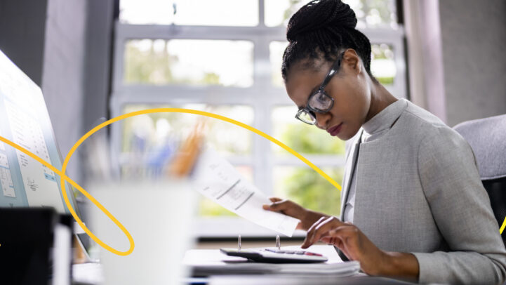 A woman wearing black glasses with her hair tied into a bun, using a calculator while sitting at a desk looking into opening a deposit account.