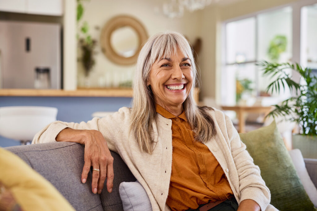 Portrait of happy retired senior woman in causal sitting on couch and smiling. Elderly healthy lady with grey hair smiling while sitting on comfortable sofa. Confident mature woman looking away with a big smile in living room at home, aspiration and fun concept.
