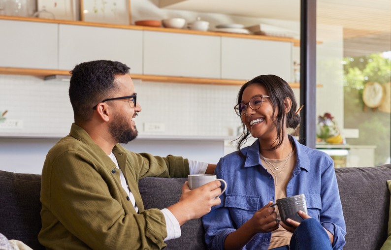 Young, mixed race couple drinking hot coffee in a mugs sitting on sofa chatting